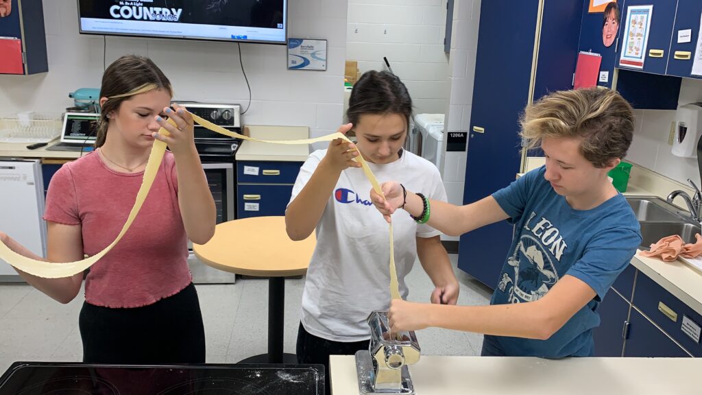 Three culinary arts students feed prepared dough to a machine to make fresh pasta.