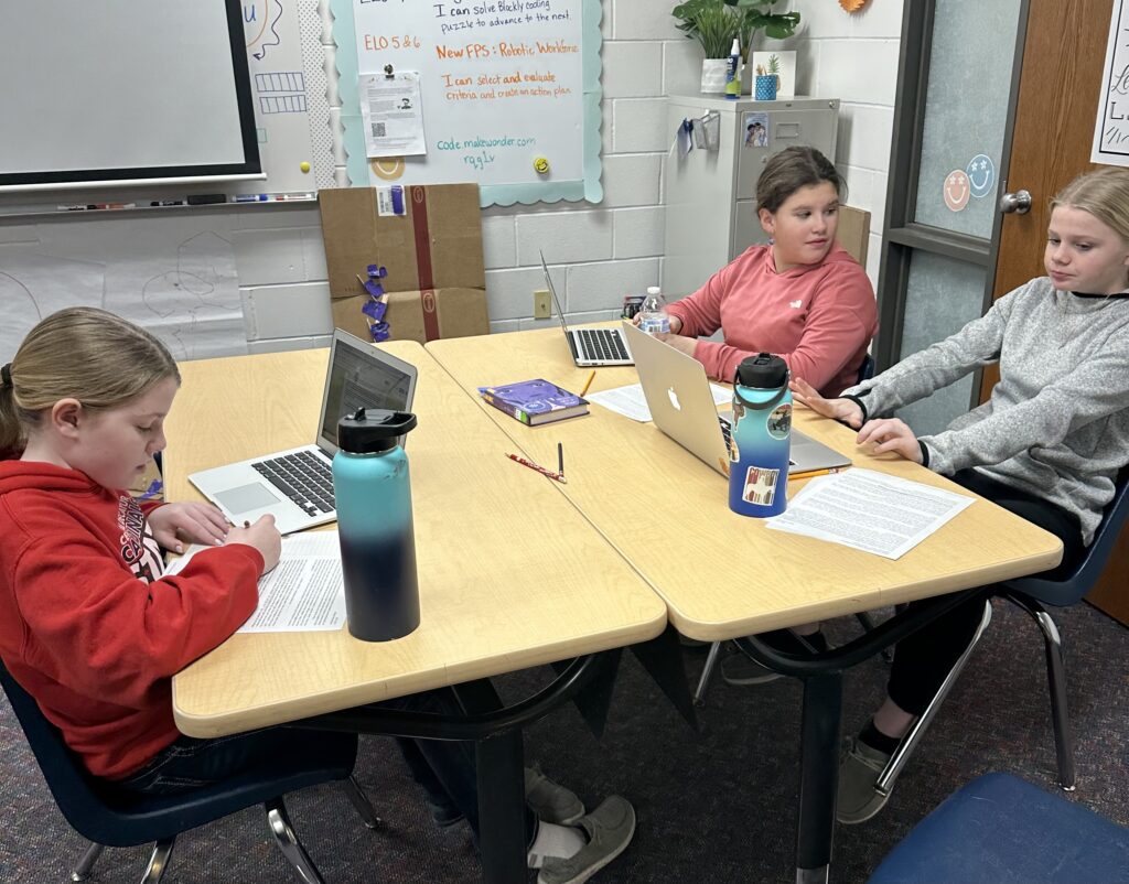 students sit at tables with laptops working to solve problems