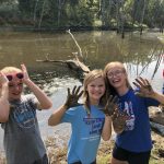 Elementary students standing in front of a lake, holding up muddy hands