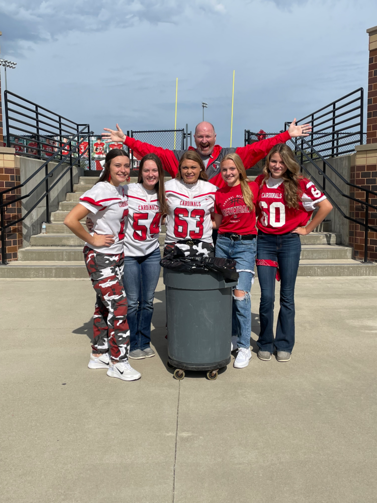 five students dressed in red and white jerseys and tshirts pose with a trash can, a teacher is standing with arms extended in the background